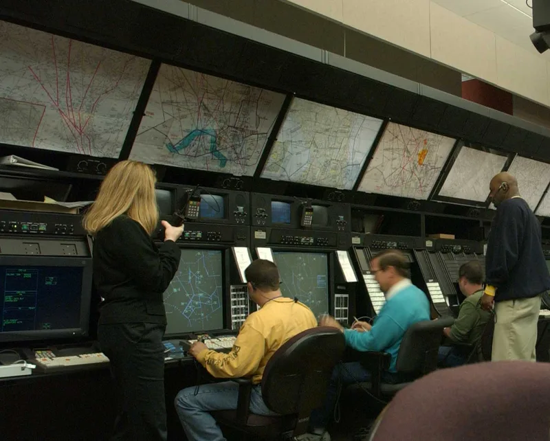 Air traffic controllers wearing headsets and microphones work at radar consoles beneath wall-mounted sector maps at an FAA air route traffic control center.