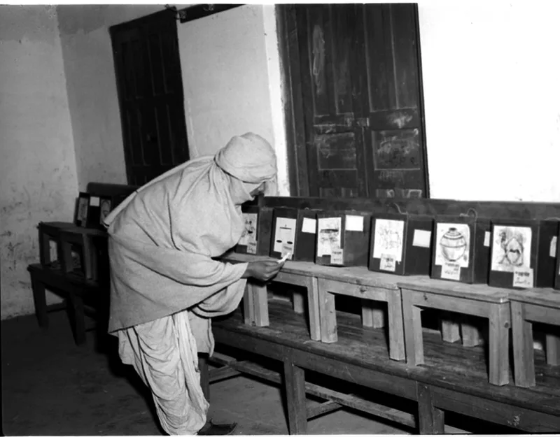 A voter in 1952 leaning over a row of separate ballot boxes, each printed with a different candidate's symbol, choosing where to post a ballot paper