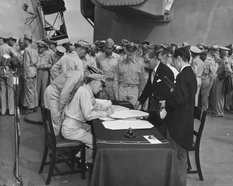 Lieutenant General Richard Sutherland leans over the signing table to correct the Japanese Instrument of Surrender, as Colonel Sidney Mashbir and Japanese Foreign Ministry official Katsuo Okazaki look on