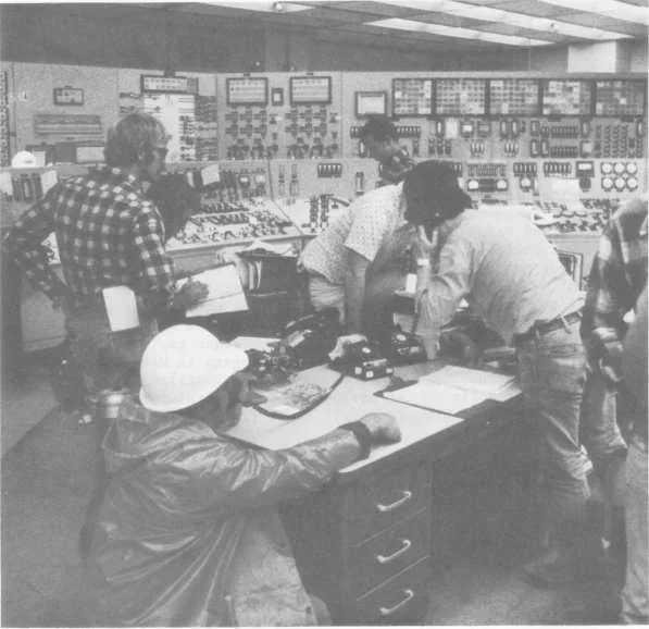 Investigators and control-room operators gathered around a desk in the TMI-2 control room days after the accident, with the instrument panel wall visible behind them.