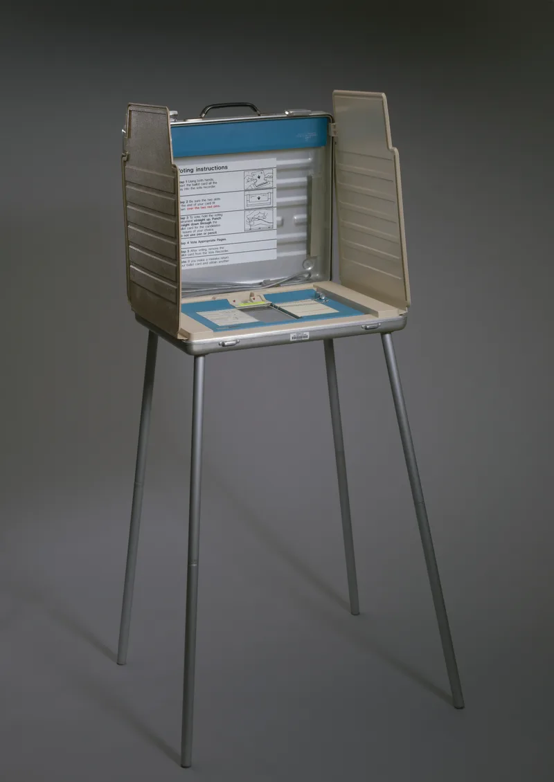 A Votomatic vote recorder mounted open on its tall folding stand, forming a free-standing voting booth, photographed against a plain grey background.